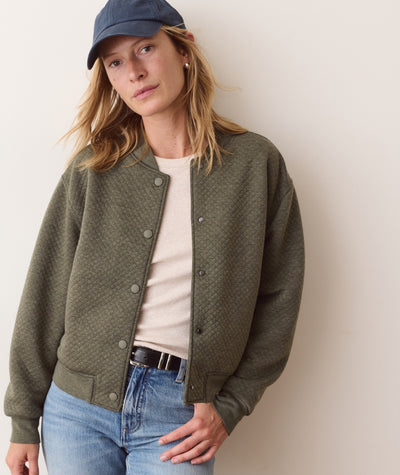 Model wearing an olive green Corbet Quilted Bomber jacket with snap buttons over a neutral top and jeans.