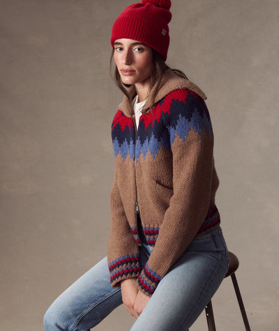 Model wearing a red Coastal Merino Pom Beanie with a pom-pom, paired with a patterned zip-up sweater.