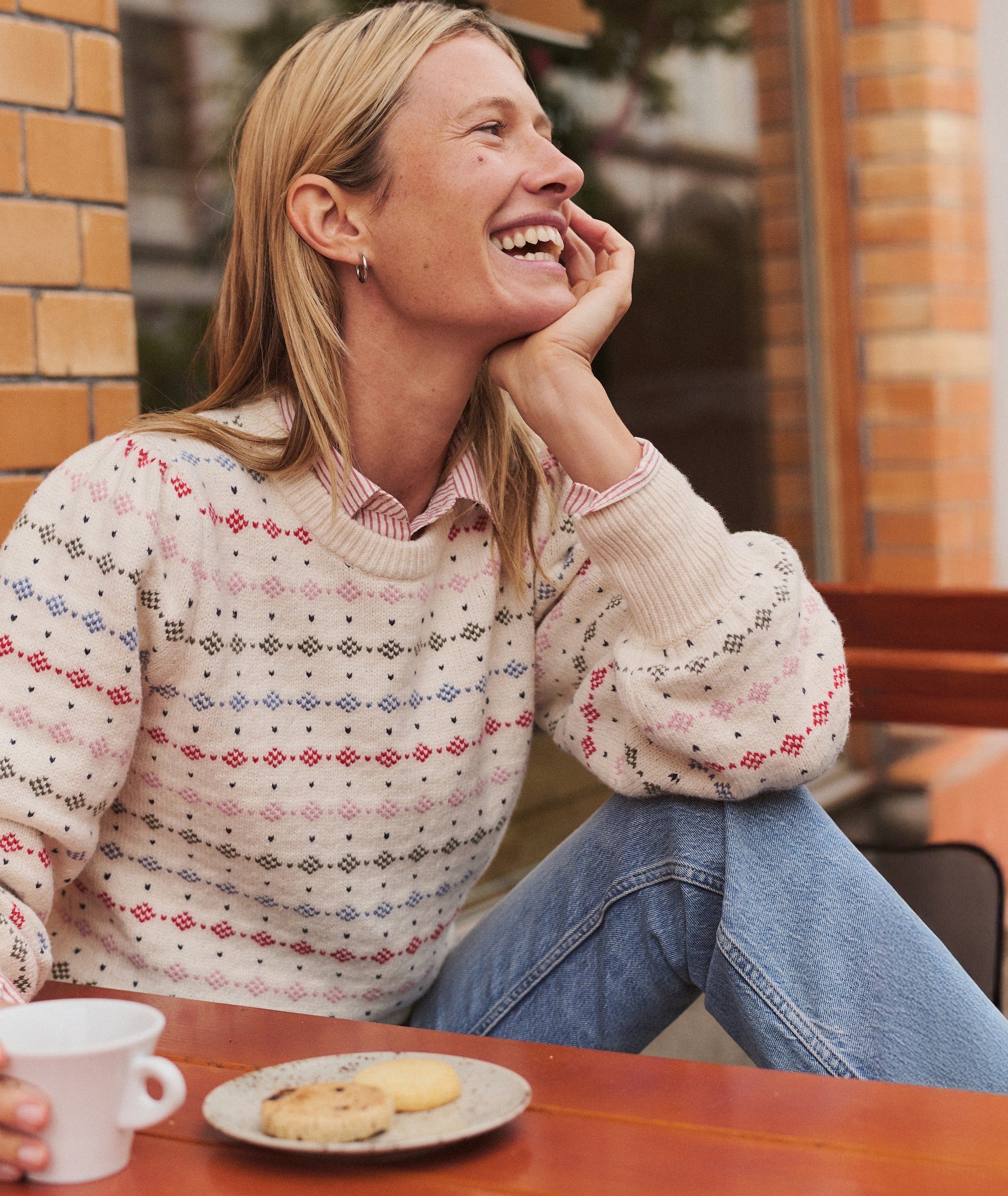 Model wearing the cream Alma Puff Sleeve Sweater with colorful fair isle patterns, seated at a cafe table.