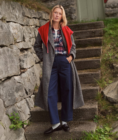 Model wears the grey herringbone Aspen Coat over a plaid shirt and blue trousers on stone steps.