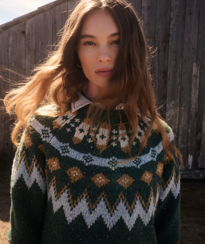 A woman wearing a dark green knit sweater with a traditional Fair Isle pattern in cream, white, and brown, standing outdoors in front of a weathered wooden wall.