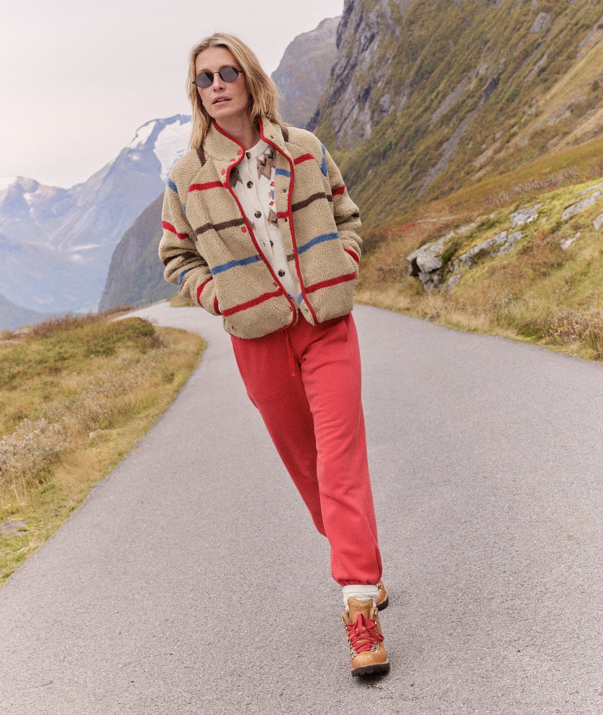 Model wears red Cloud 9 Fleece Jogger with striped fleece jacket and hiking boots on a mountain road.