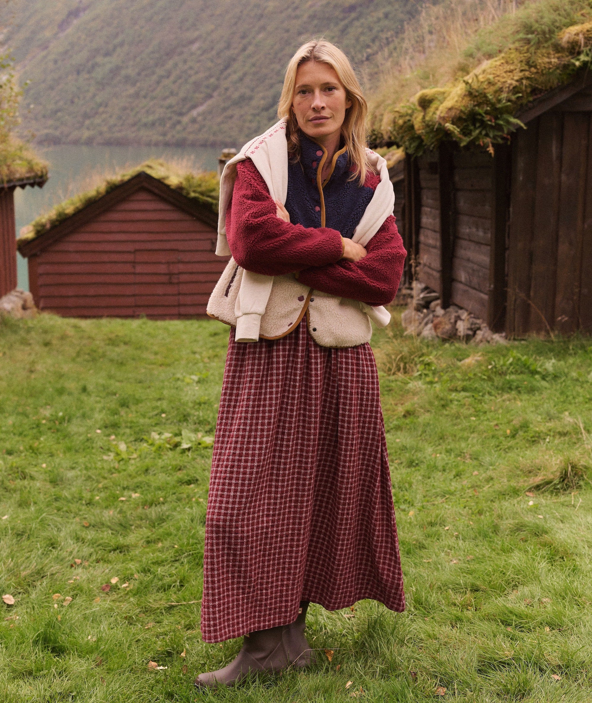 Model wearing the red plaid Gia Oxford Midi Skirt with a fleece vest and sweater in a rustic setting.